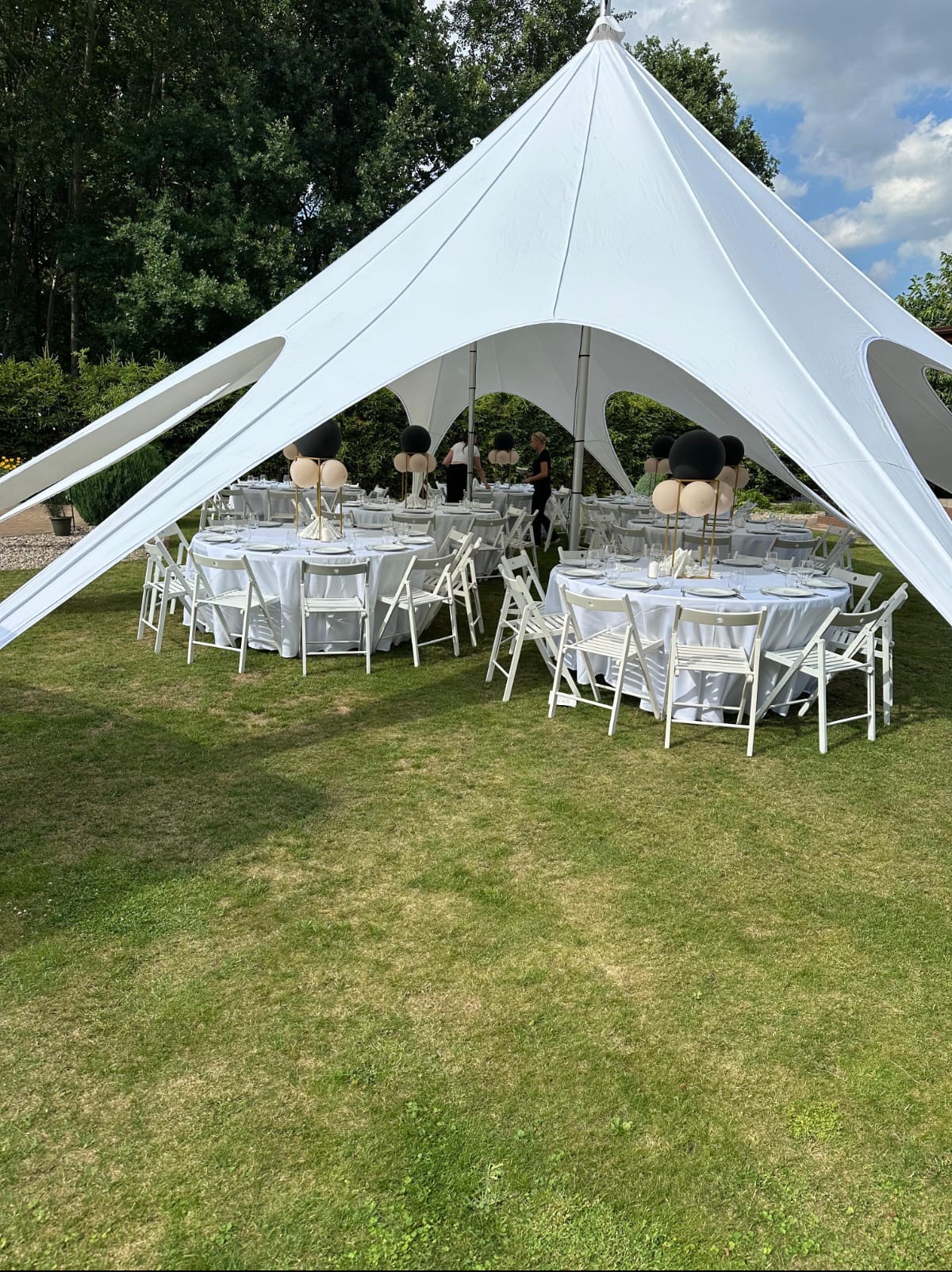 Biały namiot sferyczny na trawniku, pod nim okrągłe stoły z białymi obrusami, krzesła i dekoracje z balonów w kolorach czarnym i beżowym, przygotowane do przyjęcia.