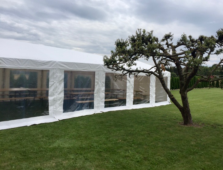 Biały namiot z przezroczystymi ścianami na zielonym trawniku, obok drzewo z powykręcanymi gałęziami, pochmurne niebo w tle.