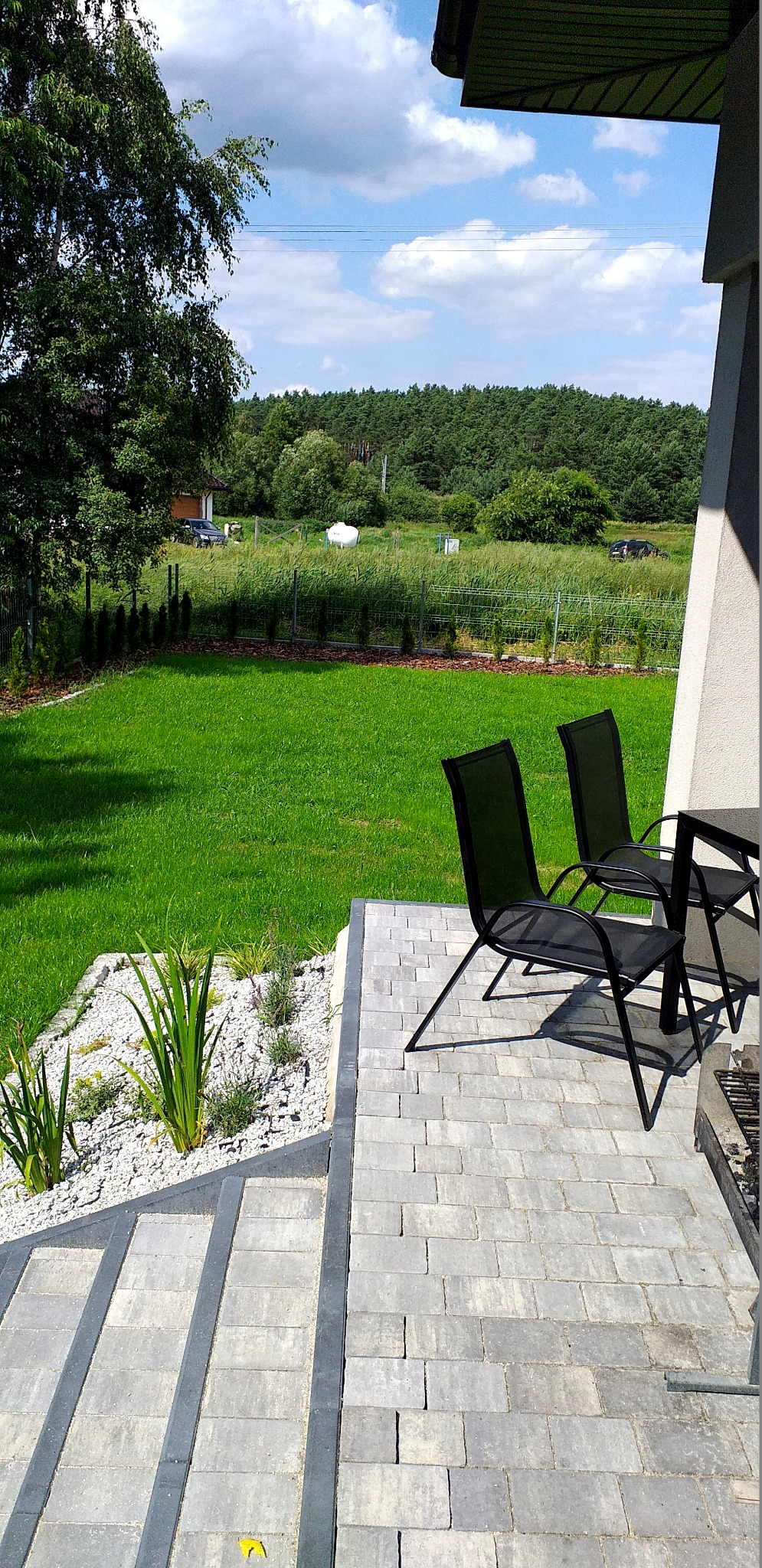 Widok na zadbany trawnik z tarasu wyłożonego szarą kostką brukową, z dwoma czarnymi krzesłami i rabatą z białym żwirem i roślinami ozdobnymi.