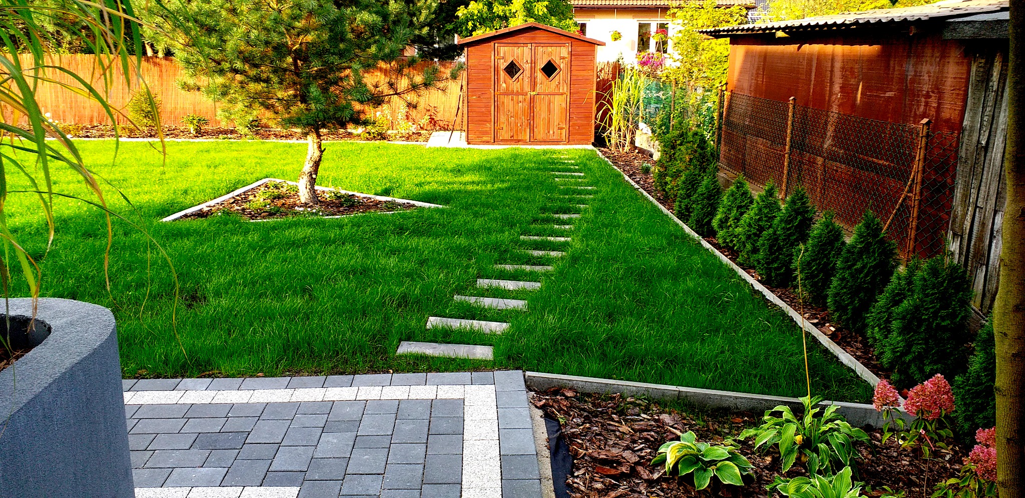 Zadbany ogród z równo przystrzyżonym trawnikiem, ścieżką z płyt betonowych prowadzącą do drewnianej szopy, obrzeżami z kostki brukowej i nasadzeniami z tui szmaragd.