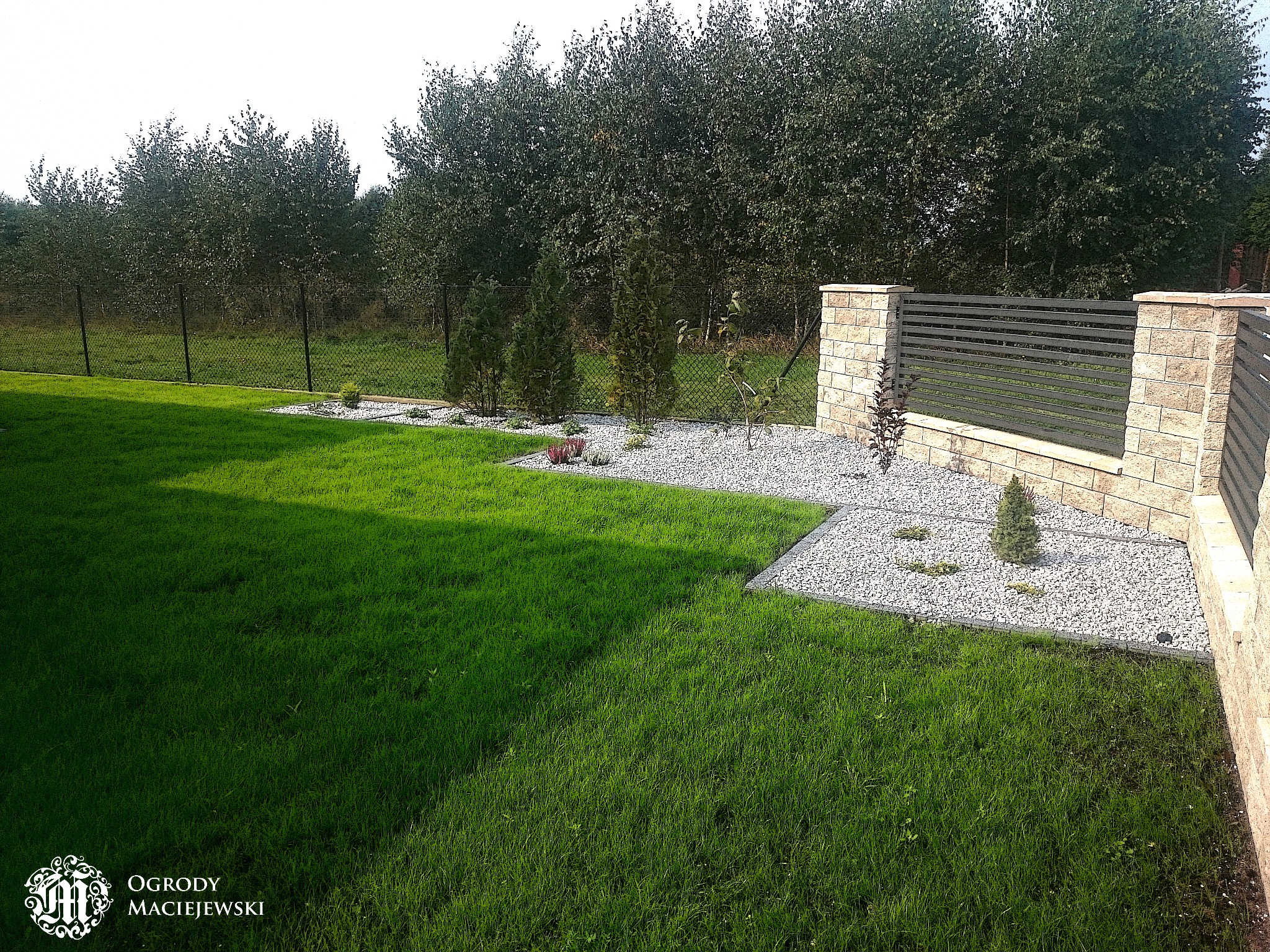 Zadbany trawnik z obrzeżami z białego grysu i niskimi nasadzeniami, w tle żywopłot z drzew liściastych i nowoczesne ogrodzenie z kamienia łupanego i metalowych przęseł.