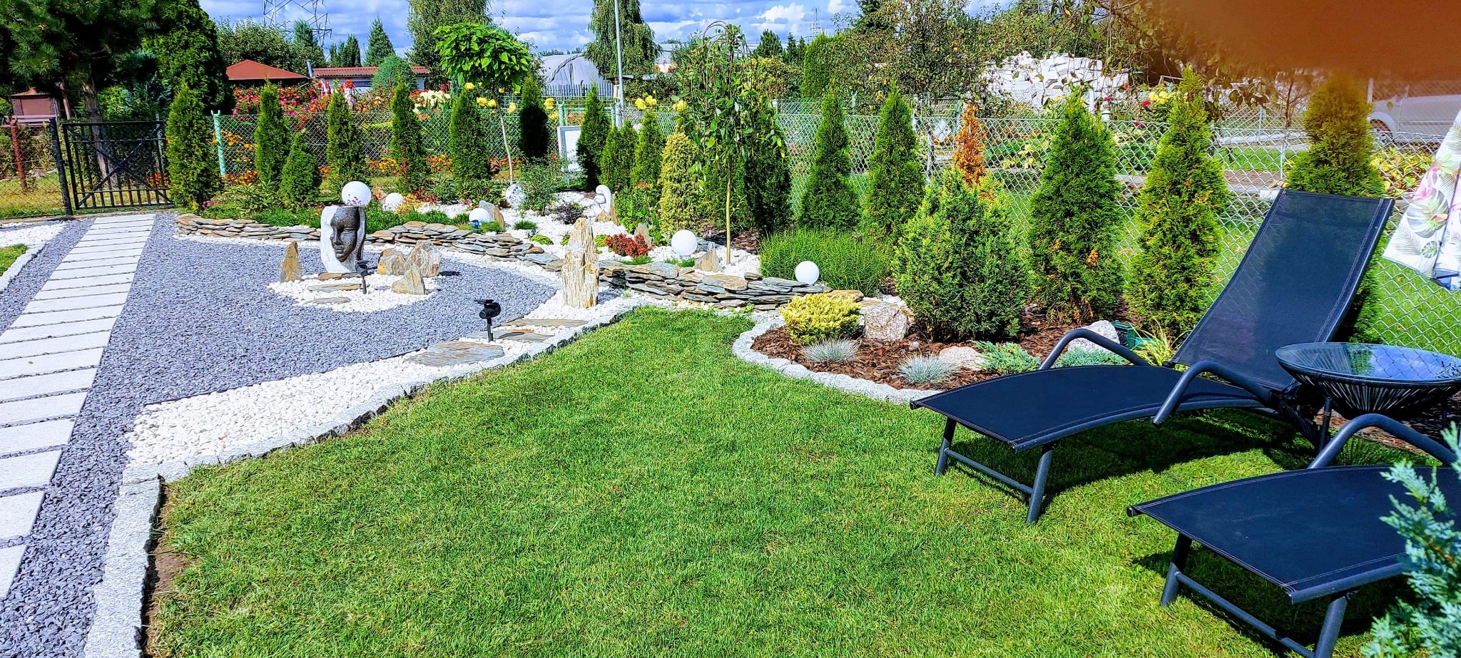 Starannie zaprojektowany ogród z równo przystrzyżonym trawnikiem, ścieżką z jasnych i ciemnych kamieni, ozdobnymi kamieniami, rzeźbą twarzy i leżakami ogrodowymi.