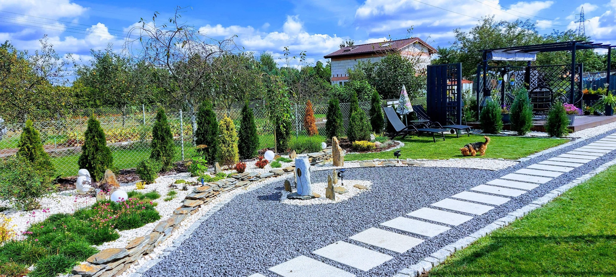 Ogród z ścieżką z płyt, żwirową rabatą z ozdobnymi kamieniami i rzeźbami, trawnikiem oraz zadaszoną konstrukcją w tle. W rogu kadru biega mały pies.