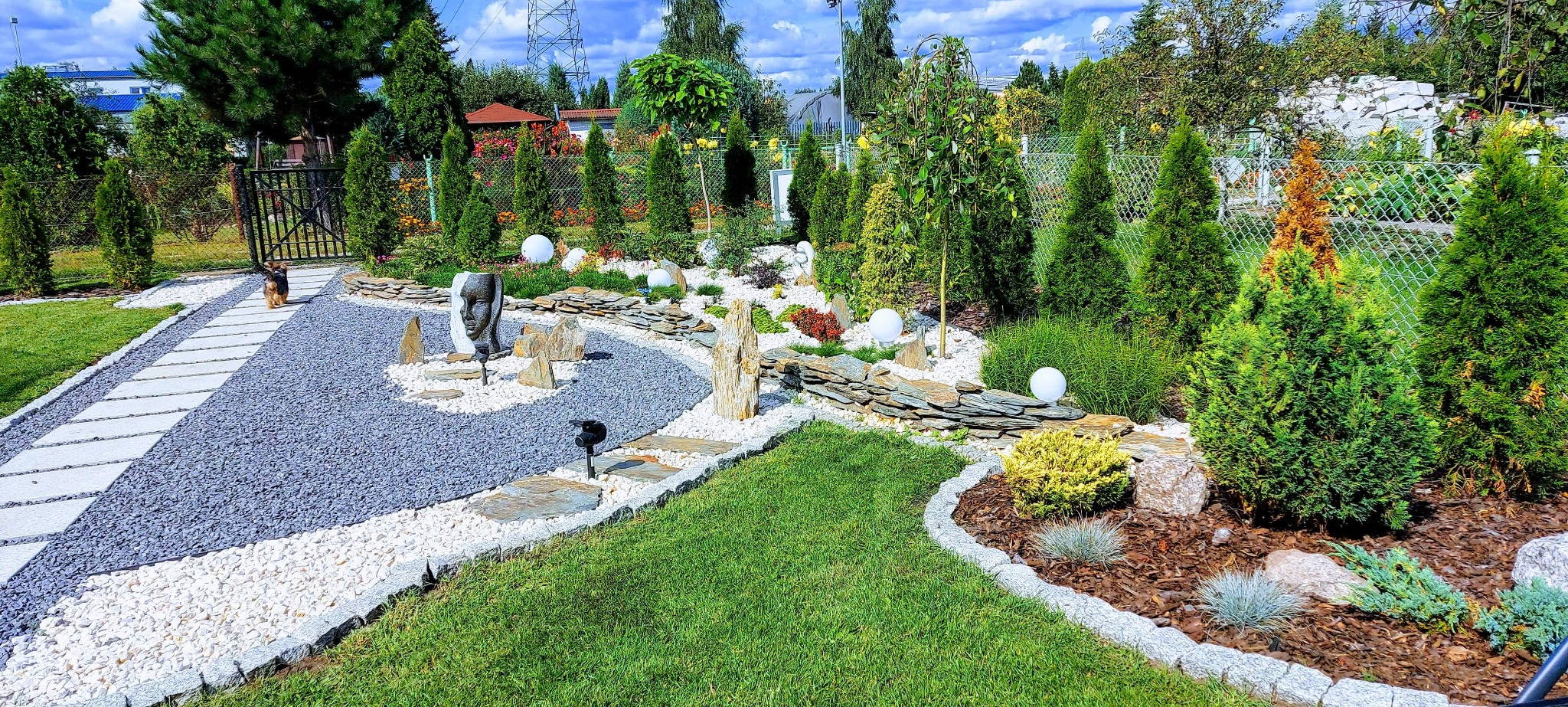 Ogród z elementami dekoracyjnymi: ścieżki z kamienia, rzeźba twarzy, białe kule jako oświetlenie, ozdobne krzewy i drzewa iglaste.