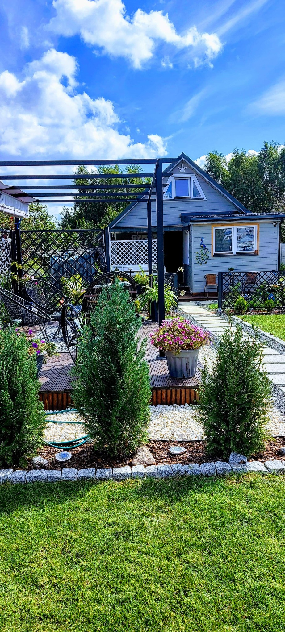 Zadbany ogród przydomowy z drewnianym tarasem, ścieżką z płyt, ozdobnymi roślinami w donicach, obrzeżami z kostki granitowej i elementami dekoracyjnymi.