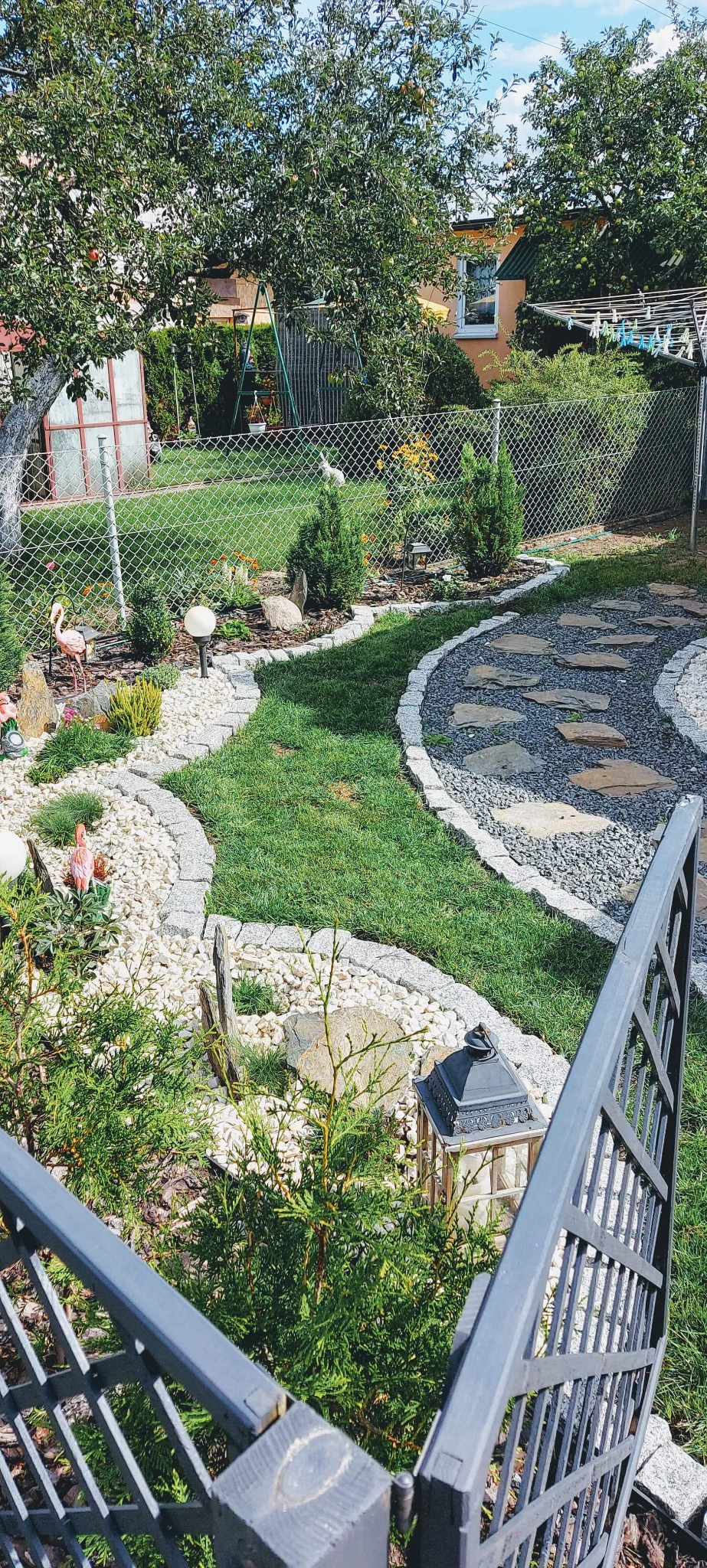 Ogród z trawnikiem, ścieżką z kamieni i żwiru, rabatami z białego kamienia i dekoracjami, widok zza metalowego ogrodzenia.