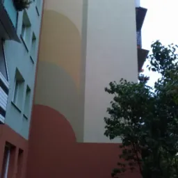 Wysoki blok po renowacji elewacji, z geometrycznym wzorem w odcieniach beżu i brązu, widoczny z dołu, z drzewem w prawym dolnym rogu.