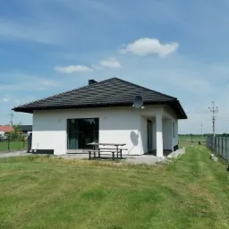 Jednopiętrowy dom z białą elewacją i ciemnym dachem z dachówki, na trawniku w słoneczny dzień, z widoczną anteną satelitarną i drewnianym stołem piknikowym.