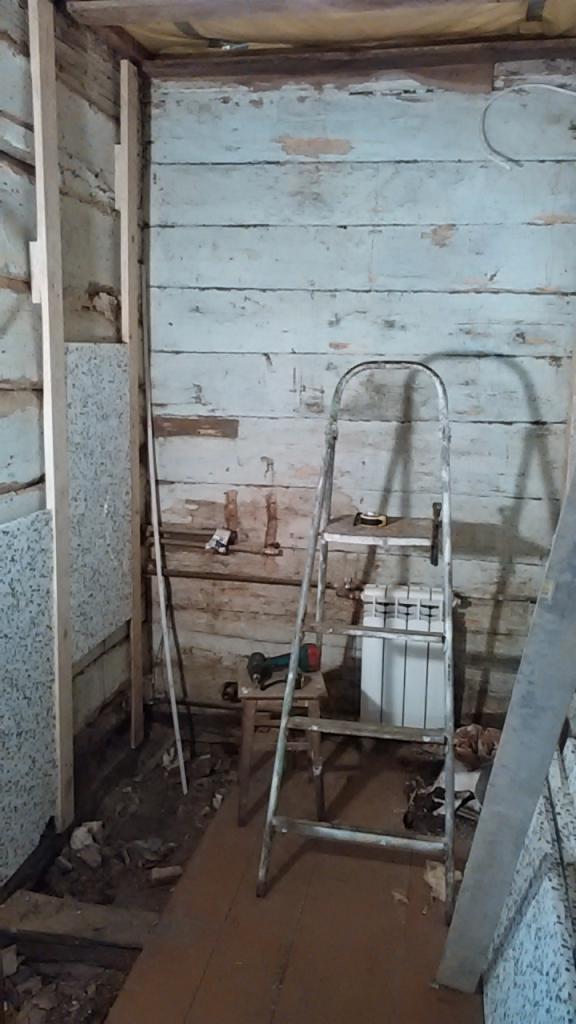 Wnętrze w trakcie remontu: ściany z drewnianą boazerią, drabina, grzejnik, fragment płyty i narzędzia. Widoczne ślady po instalacjach i przygotowania do dalszych prac.