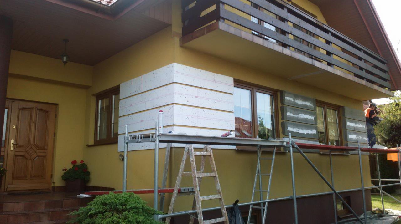Ocieplanie elewacji domu jednorodzinnego, widoczne rusztowania, częściowo nałożony styropian. Pracownik w trakcie pracy. Dom z balkonem i drewnianymi elementami.