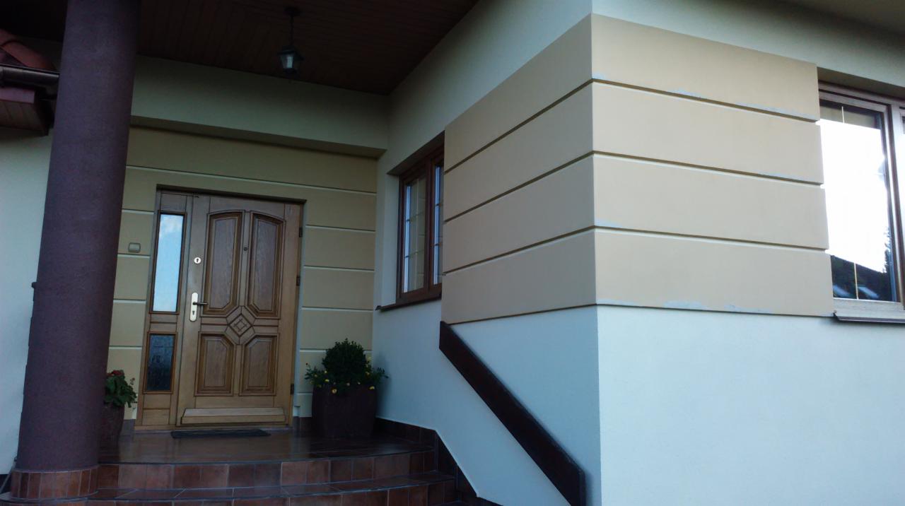 Wejście do domu po renowacji: kolumna, drewniane drzwi z szybką, jasna elewacja z poziomymi pasami w odcieniach beżu i brązu, okno z drewnianą ramą.