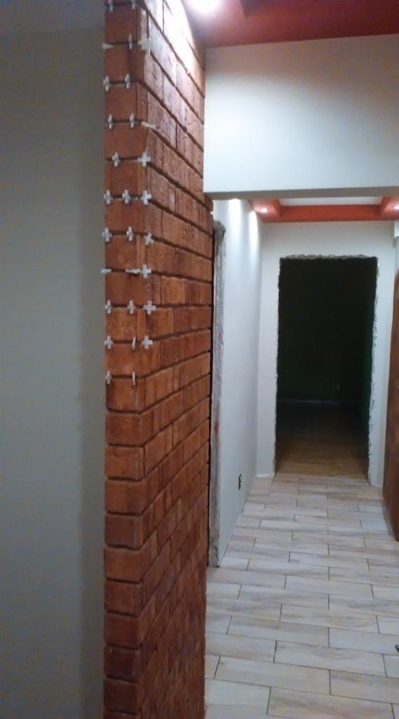 Ściana z cegły klinkierowej w trakcie montażu, widoczne krzyżyki dystansowe. Podłoga wyłożona płytkami imitującymi drewno, fragment korytarza z oświetleniem.