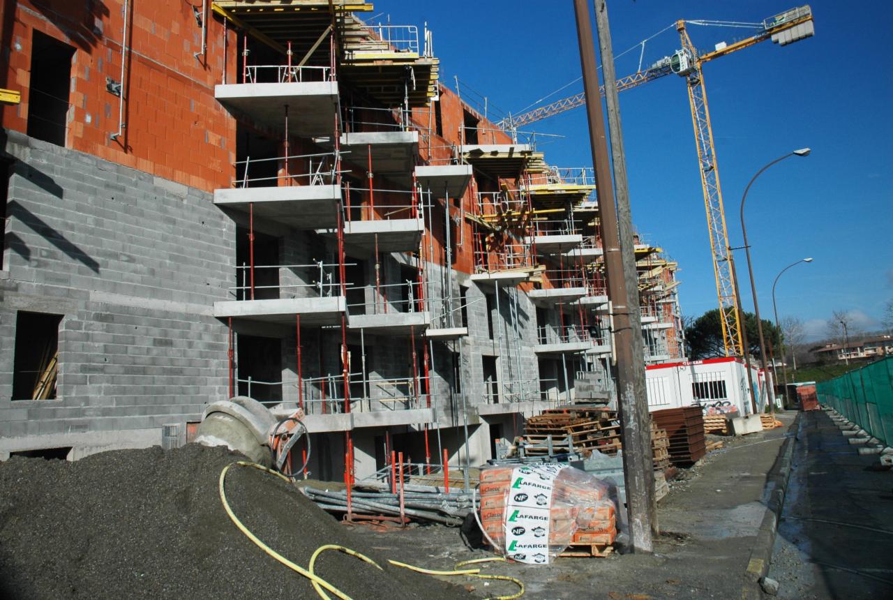 Wielorodzinny budynek mieszkalny w trakcie budowy, widoczne rusztowania, dźwig budowlany, mieszalnik do betonu i materiały budowlane Lafarge.