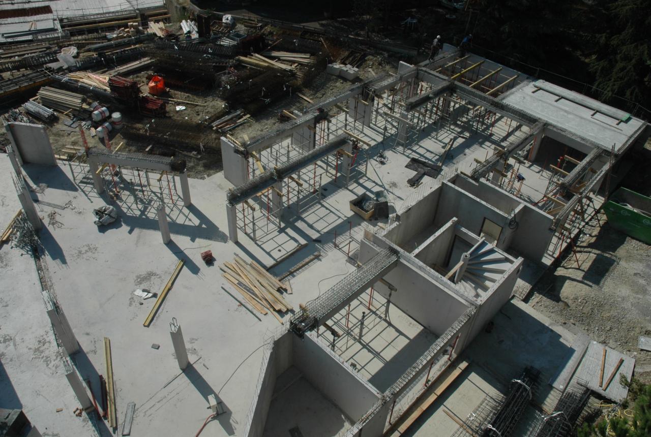 Wysoki kąt widzenia na budowę budynku mieszkalnego wielorodzinnego w trakcie wznoszenia ścian i stropów, widoczne zbrojenia, szalunki, składowane materiały budowlane i częściowo wykonana klatka...