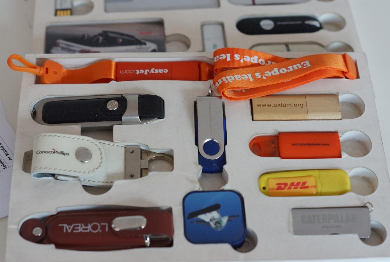 Zestaw promocyjnych pendrive'ów z logo różnych firm (EasyJet, Oxfam, DHL, L'Oreal, ConocoPhillips, Caterpillar, New Balance) umieszczonych w pudełku z przegródkami.