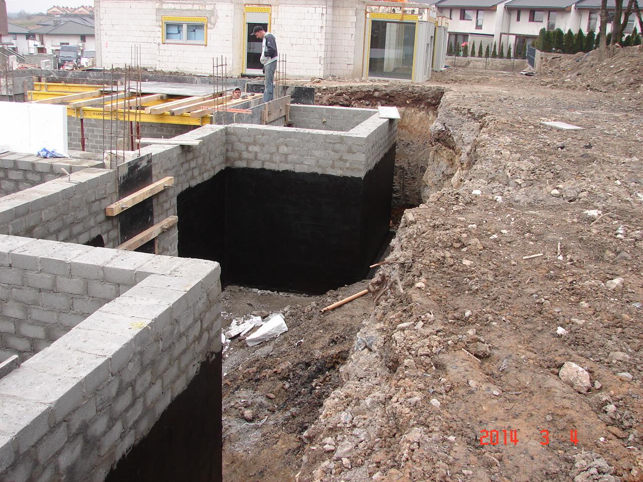 Fundamenty domu w trakcie budowy: ściany z bloczków, izolacja przeciwwilgociowa, wykop. Widoczni pracownicy i materiały budowlane. Domy w tle.