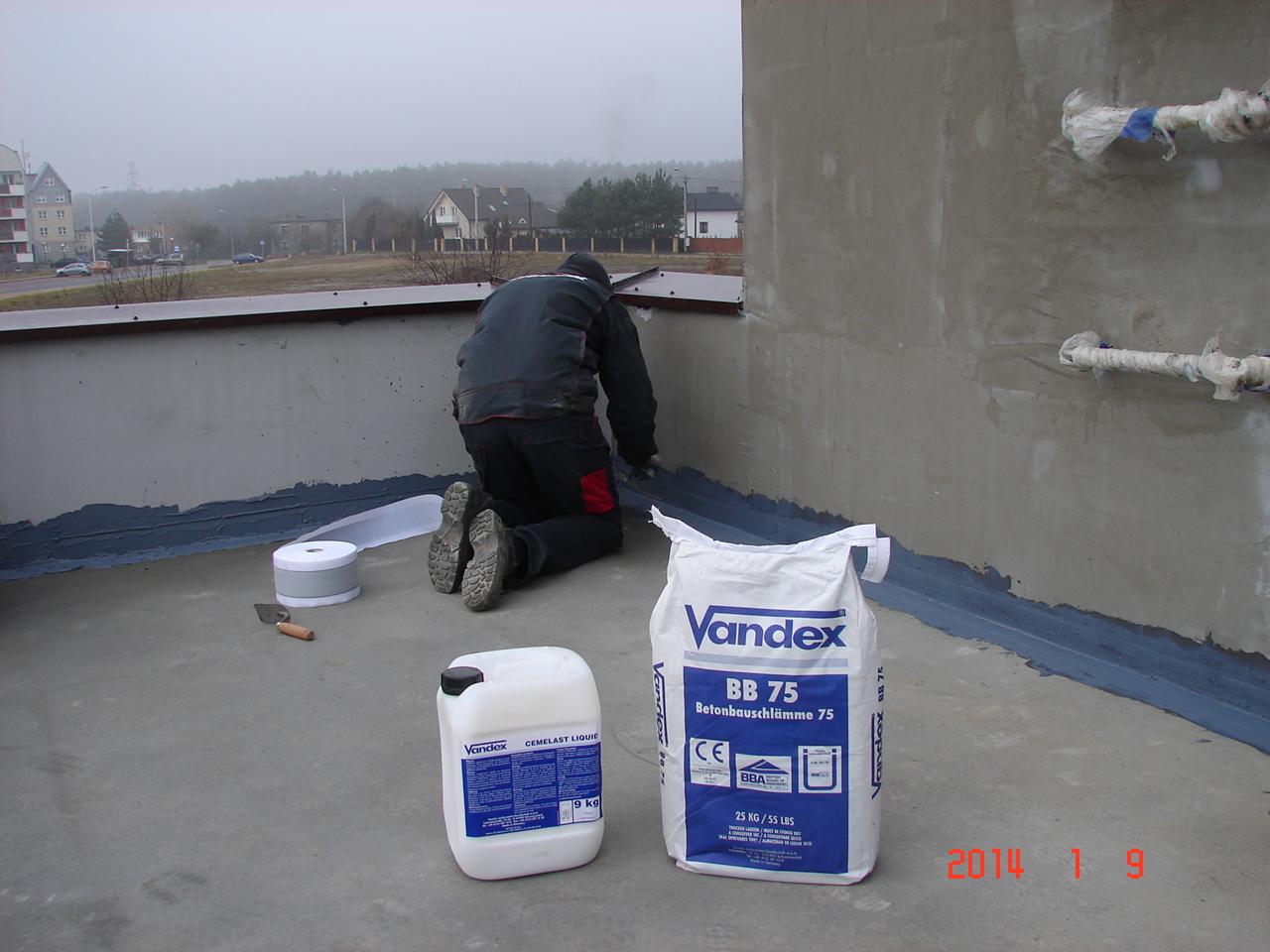 Hydroizolacja dachu płaskiego: pracownik aplikuje masę uszczelniającą Vandex BB 75. Widoczne opakowania produktu, taśma i kielnia. W tle zabudowa miejska w pochmurny dzień.