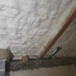 Poddasze ocieplone białą pianką poliuretanową, widoczne drewniane elementy konstrukcyjne i fragment ściany z szarego betonu komórkowego.