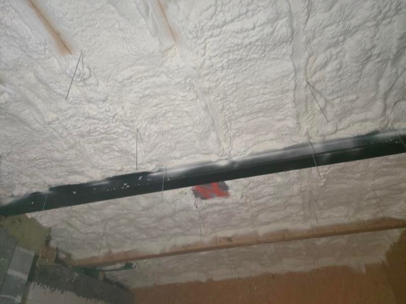 Świeżo nałożona, nierówna warstwa jasnej pianki izolacyjnej na suficie z widocznymi elementami konstrukcyjnymi, w tym czarną rurą i drewnianymi belkami.