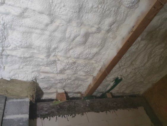 Poddasze ocieplone białą pianką poliuretanową, widoczne drewniane elementy konstrukcyjne i fragment ściany z szarego betonu komórkowego.