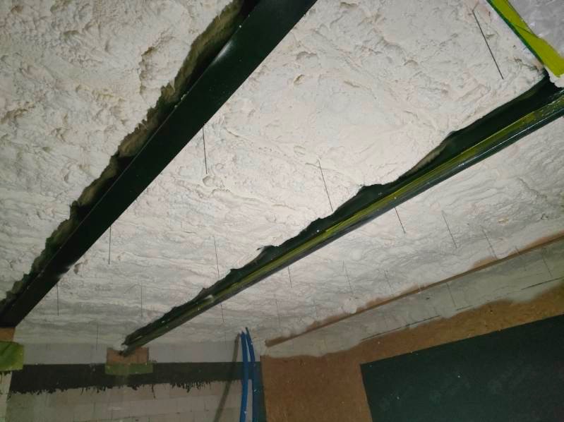 Biała pianka poliuretanowa natryśnięta na strop między metalowymi belkami, widoczne wystające cienkie druty mocujące, fragment ściany z płytami OSB.