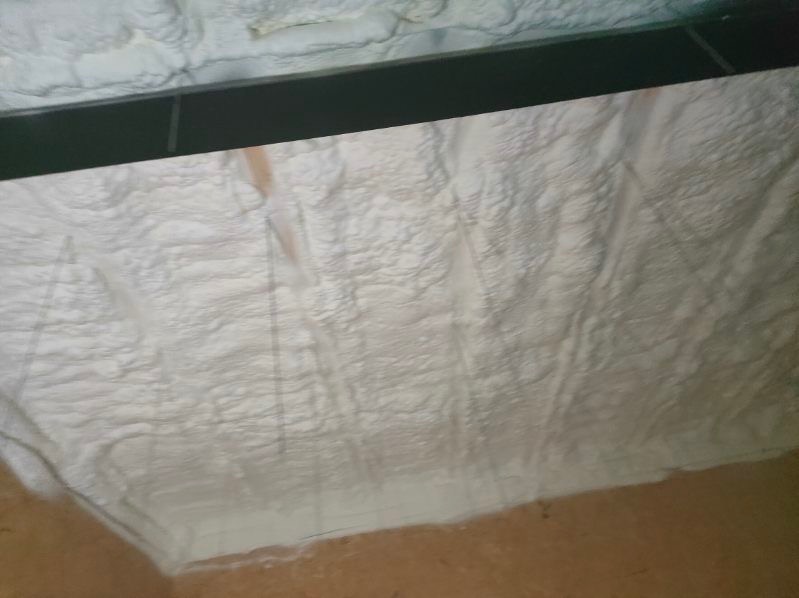 Świeżo nałożona, nierówna warstwa białej pianki poliuretanowej na ścianie wewnętrznej pod stropem, widoczny ciemny element konstrukcyjny.