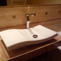 Biała, designerska umywalka z chromowaną baterią na blacie z płytek imitujących marmur. W tle ściana wyłożona płytkami i fragment wanny.