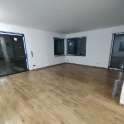 Jasne, drewnopodobne panele ułożone w dużym, pustym pokoju z białymi ścianami i oknami zabezpieczonymi folią malarską.