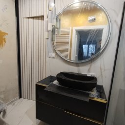 Brzuszkiewicz Arek - Nowoczesna łazienka w trakcie wykończenia: czarna umywalka nablatowa na szafce z złotymi akcentami, okrągłe lustro z podświetleniem LED, dekoracyjne lamele ścienne i marmurowe płytki.