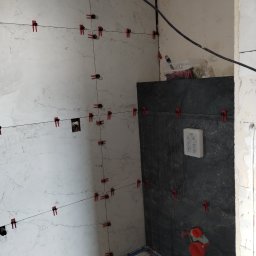 Brzuszkiewicz Arek - Układanie płytek imitujących marmur na ścianie, z użyciem klipsów poziomujących i krzyżyków dystansowych, widoczne przyłącze wodne i fragment ściany z ciemnymi płytkami.