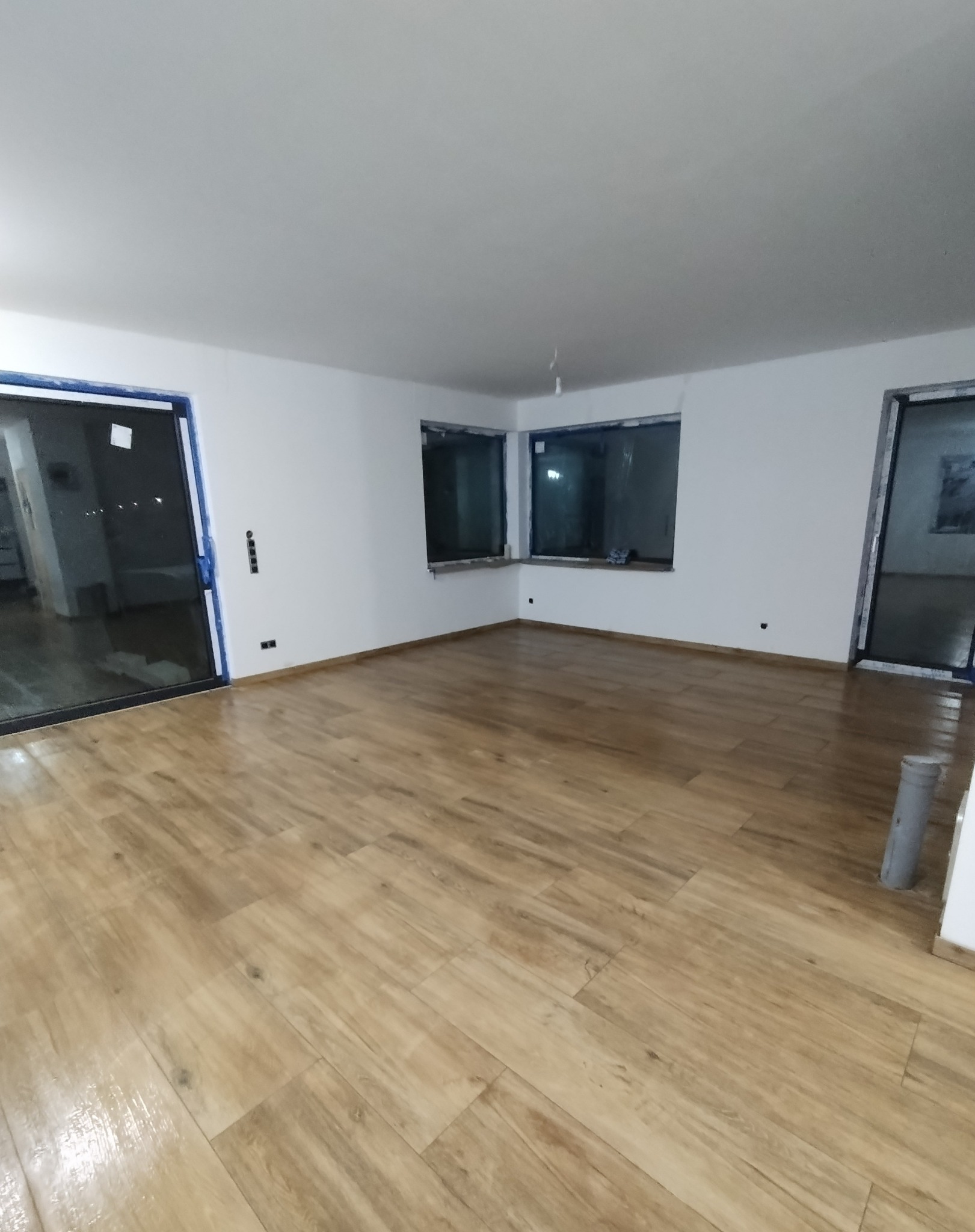 Jasne, drewnopodobne panele ułożone w dużym, pustym pokoju z białymi ścianami i oknami zabezpieczonymi folią malarską.