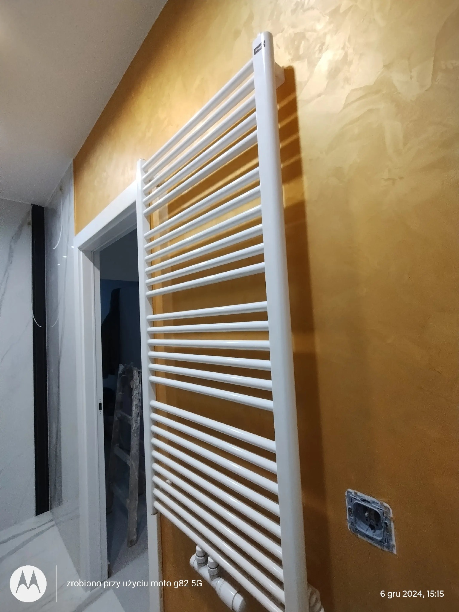 Biały, drabinkowy grzejnik łazienkowy zamontowany na złotej ścianie z efektem dekoracyjnym, widoczny fragment niezabezpieczonego gniazdka elektrycznego i drabina w tle.