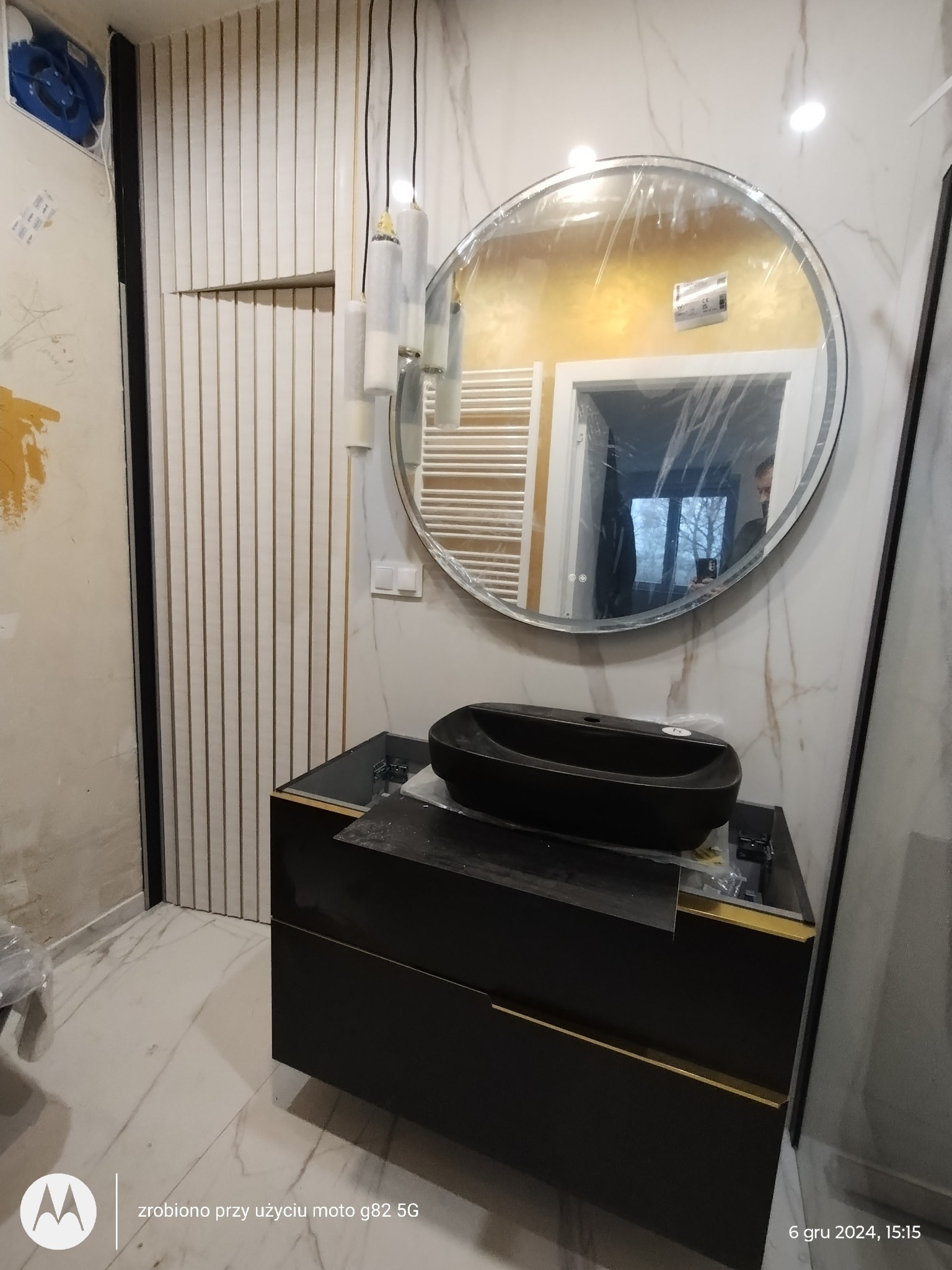 Nowoczesna łazienka w trakcie wykończenia: czarna umywalka nablatowa na szafce z złotymi akcentami, okrągłe lustro z podświetleniem LED, dekoracyjne lamele ścienne i marmurowe płytki.