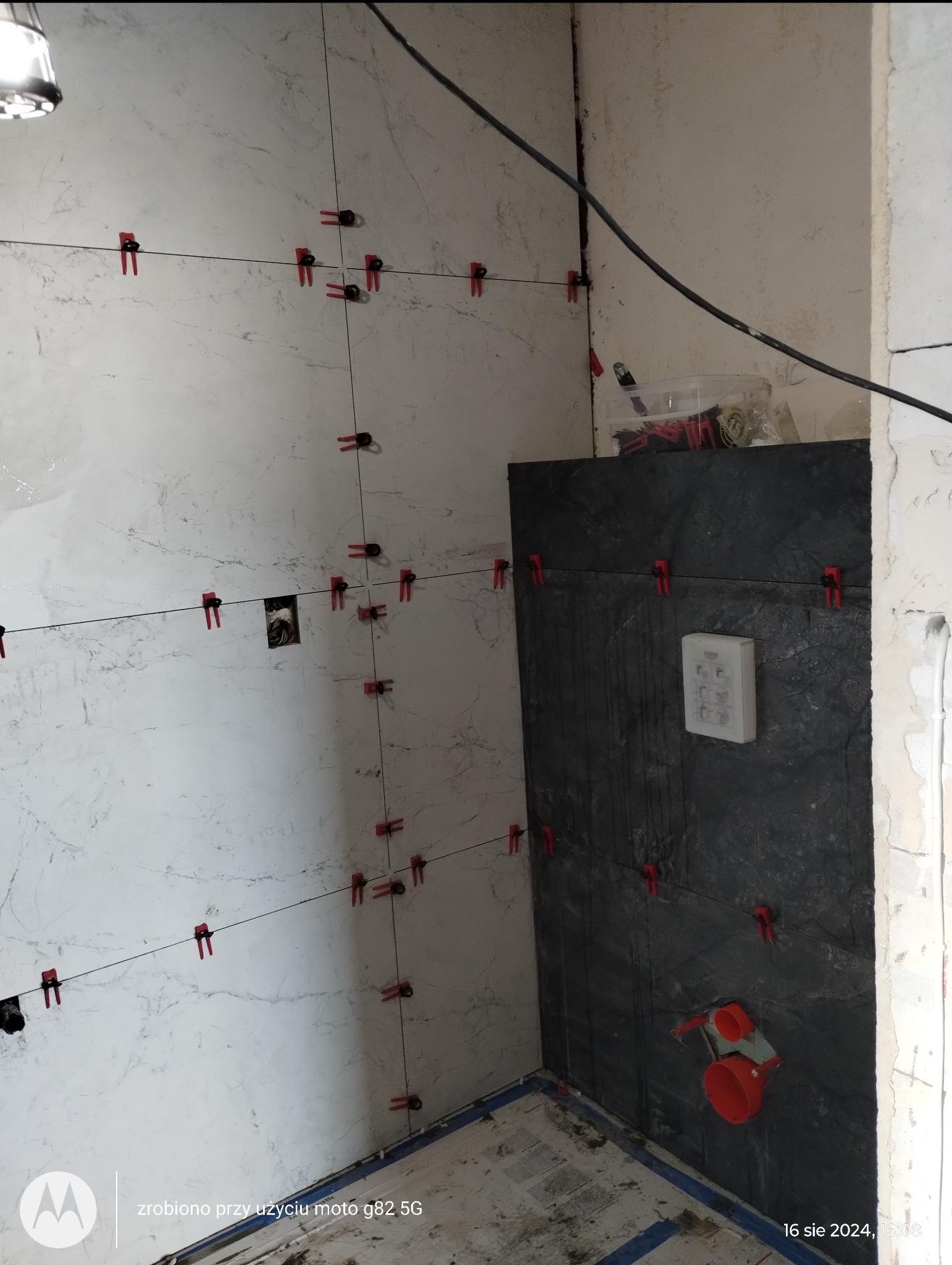 Układanie płytek imitujących marmur na ścianie, z użyciem klipsów poziomujących i krzyżyków dystansowych, widoczne przyłącze wodne i fragment ściany z ciemnymi płytkami.