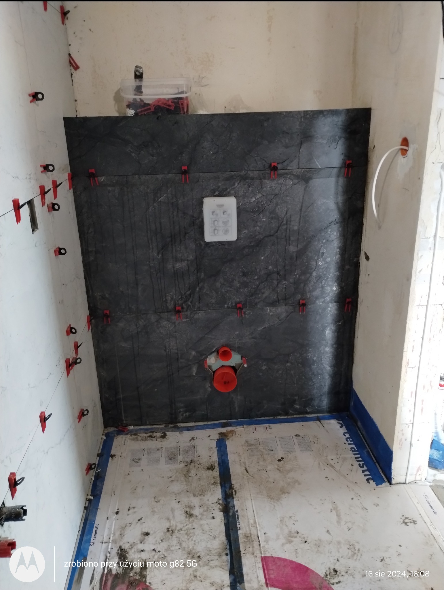 Montaż czarnych płytek imitujących kamień na ścianie w łazience, widoczne czerwone klipsy poziomujące, instalacja podtynkowego WC i fragment instalacji wodnej, podłoga zabezpieczona kartonem.