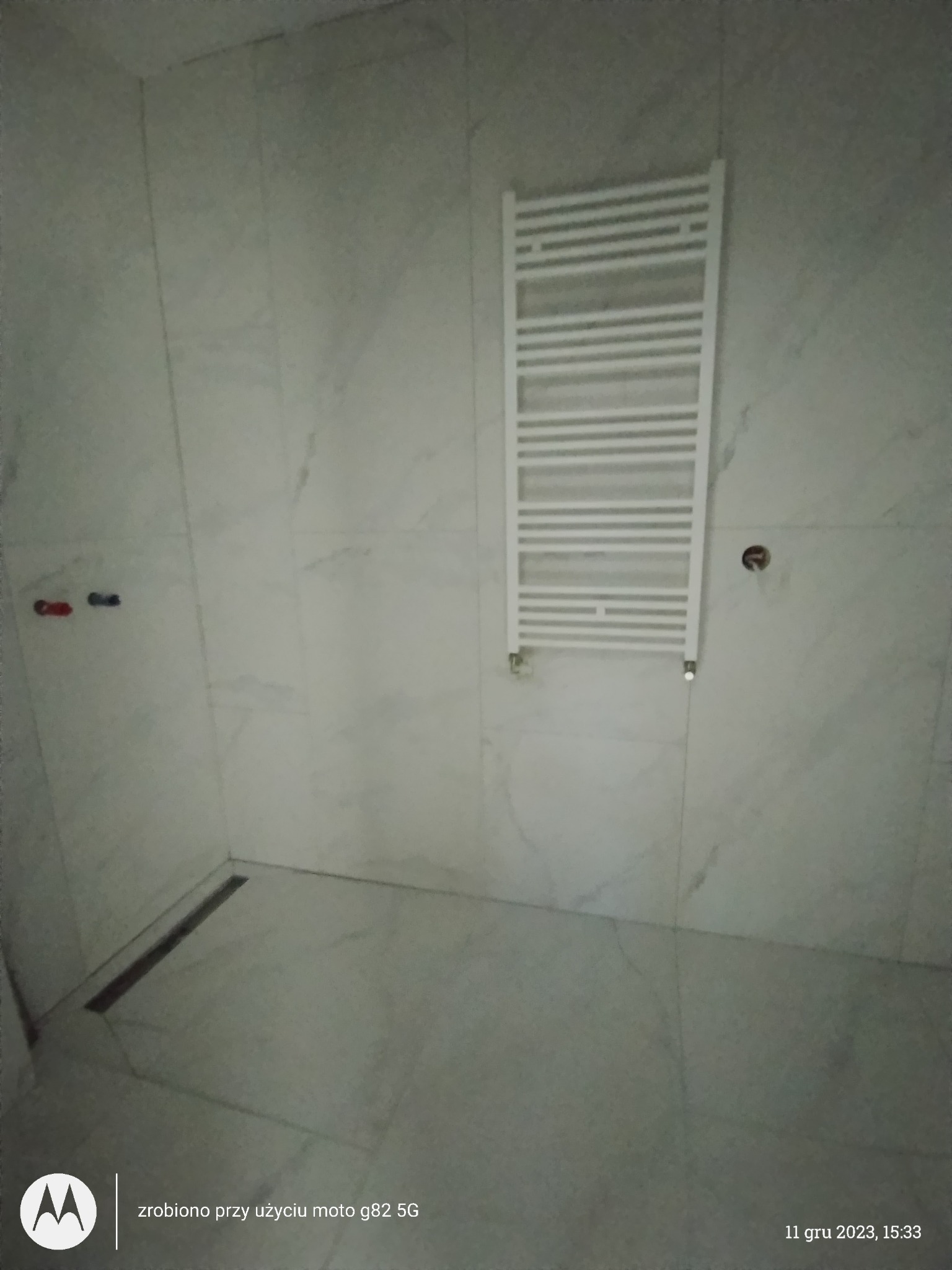 Wykończona łazienka z białymi marmurkowymi płytkami, odpływem liniowym w podłodze i białym grzejnikiem drabinkowym na ścianie.