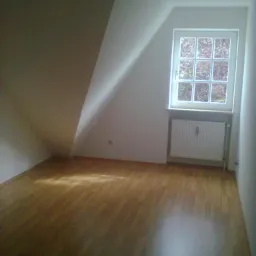 Poddasze po remoncie: drewniana podłoga, białe ściany, okno z widokiem na zewnątrz. Skos dachu z oknem doświetla wnętrze. Grzejnik pod oknem.