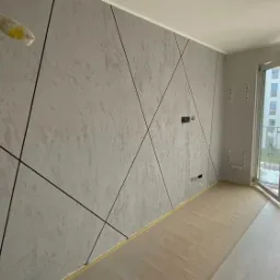 Ściana w pokoju po nałożeniu dekoracyjnej szarej gładzi z czarnymi liniami tworzącymi geometryczny wzór, zabezpieczona podłoga folią malarską, widok na balkon i budynki za oknem.