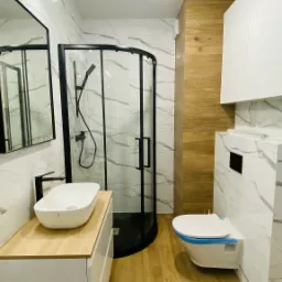 Nowoczesna łazienka po remoncie: kabina prysznicowa z czarną ramą, umywalka nablatowa na drewnianym blacie, toaleta podwieszana z niebieską folią zabezpieczającą, marmurowe płytki na ścianach...