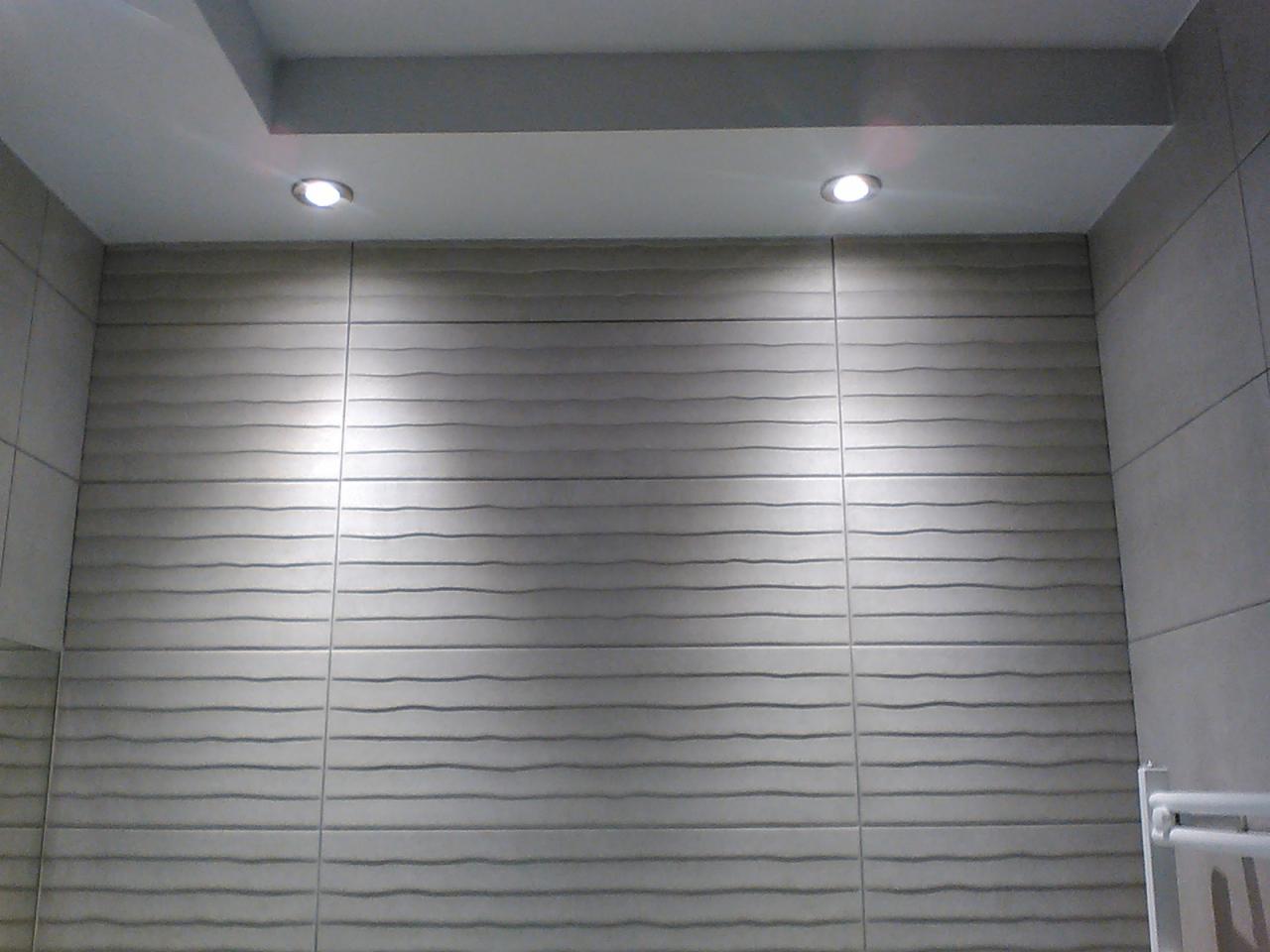 Ściana łazienki wyłożona płytkami z falującym wzorem, oświetlona dwoma punktowymi lampami w suficie podwieszanym.