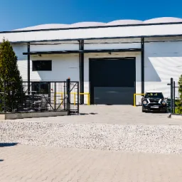 Nowoczesna hala magazynowa z białymi ścianami i charakterystycznym zaokrąglonym dachem, duża brama wjazdowa, ogrodzenie z siatki, zadbana zieleń i zaparkowany czarny Mini Cooper z przodu.