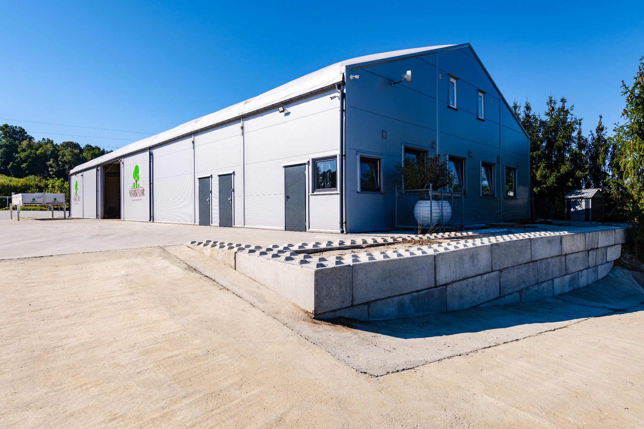 Gotowa hala w konstrukcji stalowej, z widocznym logo firmy na elewacji, betonowy murek oporowy, błękitne niebo w tle.