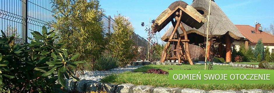 Ogród z altaną krytą strzechą i ozdobnymi kamieniami, obok zielone tuje i nowoczesne ogrodzenie panelowe. Całość w słoneczny dzień.