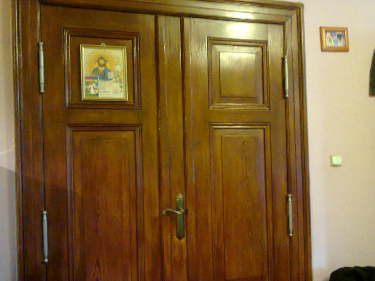 Lakierowane, dwuskrzydłowe drzwi z ramkami, zdobione obrazkiem i długimi uchwytami, na tle jasnoróżowej ściany z małym obrazkiem i włącznikiem.