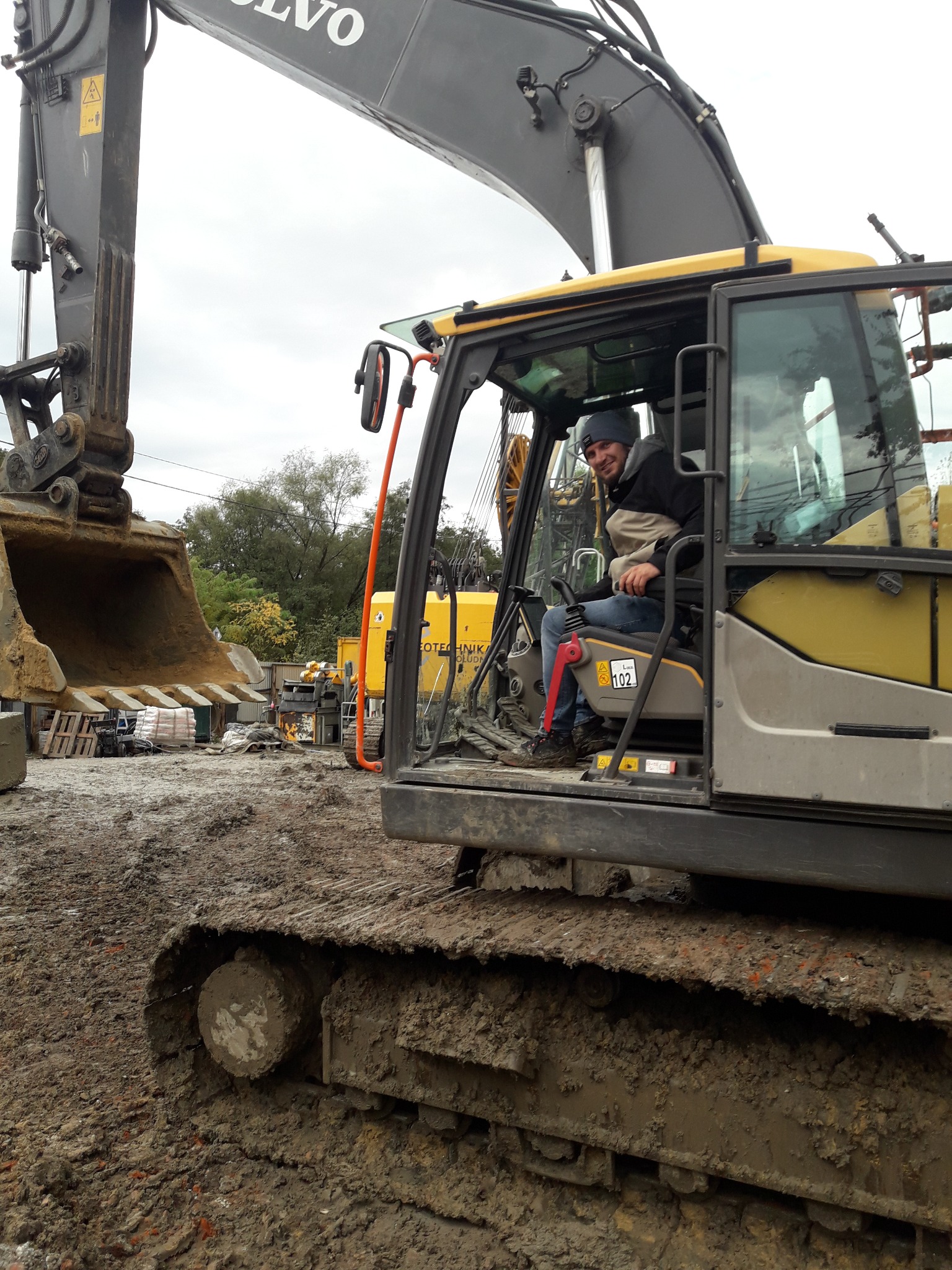 Operator w kabinie żółtej koparki Volvo, widoczny fragment łyżki i błotniste podłoże. Uśmiechnięty mężczyzna w czapce siedzi za sterami maszyny.