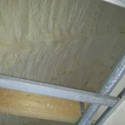 Jasna, nierówna warstwa izolacyjna z pianki natryskowej, widoczna od spodu, przylegająca do konstrukcji metalowego stelażu sufitu podwieszanego i częściowo drewnianej belki stropowej.