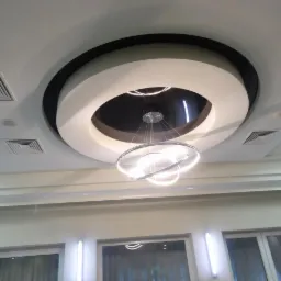 Biały sufit podwieszany z okrągłym elementem dekoracyjnym i nowoczesną lampą wiszącą z trzema okręgami LED, widoczne kratki wentylacyjne i okna z zasłonami.