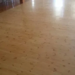 Jasne, świeżo wycyklinowane drewniane deski podłogowe z widocznymi słupami podtrzymującymi strop w tle sali.