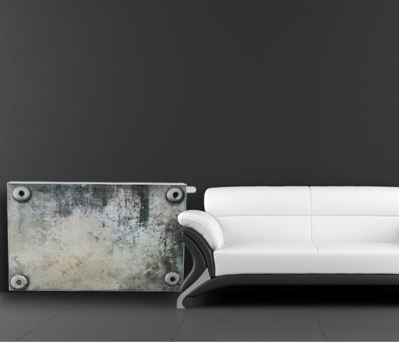 Stylowy, industrialny grzejnik żeliwny w surowym wykończeniu, z widocznymi śladami użytkowania, ustawiony obok nowoczesnej, białej sofy na tle szarej ściany.