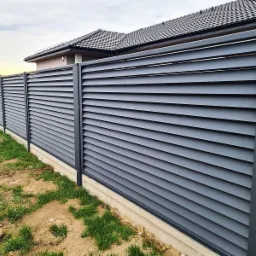 Nowoczesne, poziome ogrodzenie lamelowe w kolorze antracytowym, z widocznym betonowym podmurówką i fragmentem domu z ciemną dachówką w tle.